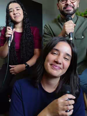 The cover image shows three people with microphones. Two women and one man are in frame. The woman in the front is smiling at the camera and holding a microphone. The woman in the back left corner is smiling at the camera and holding a microphone. The man in the back right corner is smiling at the camera and holding a microphone. The background is a plain wall and an unidentifiable setting.