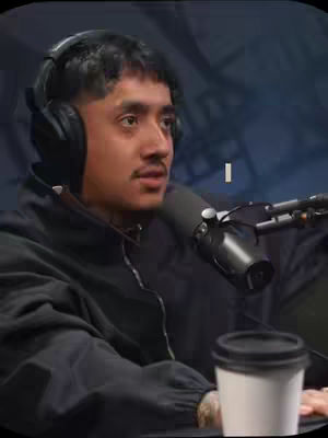 A man with dark hair and a mustache is seated, wearing a black jacket and headphones. He's holding a microphone in front of him, with a coffee cup in the foreground. The background is a blurry mix of dark colors.