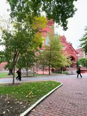 An outdoor shot with a brick path and a red brick building with arched openings in the background. Trees with green and yellowing leaves are in the foreground, and a person walks along the path. The day is overcast. 