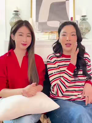 Two women are seated in a well-lit room, likely a living room, with a neutral background. The woman on the left is holding a white pillow. The woman on the right has her hand to her face. The woman on the right wears a red and white striped long-sleeved shirt and jeans and the other is wearing a red top and both are smiling. There is a painting and candles.