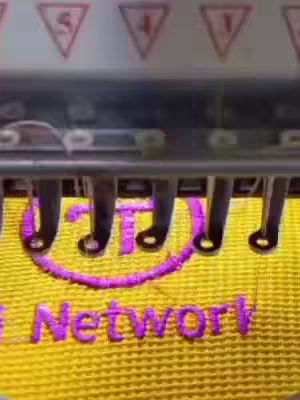 The image captures a close-up view of an embroidery machine in action, focusing on the Pi Network logo being stitched onto a yellow fabric. The text 'Pi Network' is visible, stitched in purple, and the background is blurred. The machine's needles and mechanisms are in the foreground, suggesting a process-oriented theme.
