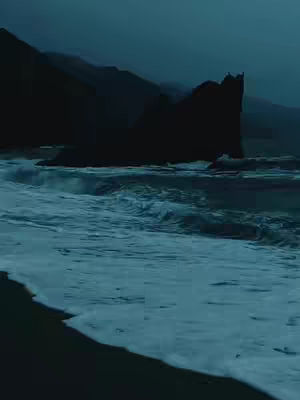 A moody, dark teal-toned image of a shoreline. The foreground shows foamy, white-crested waves washing onto a dark, sandy beach. Behind the waves, there's a silhouette of a large rock structure with jagged edges, and in the distance, there are shadowy mountains. The sky above is overcast, adding to the overall sense of depth and tranquility.