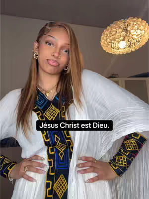 A woman with brown hair and makeup poses for the camera. She is wearing a white dress with a colorful embroidered pattern down the front and on her sleeves. Her hands are up close to her chest at the forearms. The background is neutral-colored, with a round chandelier in the upper right corner and a white wall.