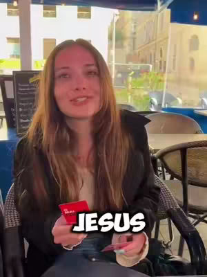 A long-haired woman, seated outdoors at a cafe table, looks at the camera with a slight smile. She holds two red cards, one with text, in front of her against a blurred background of buildings and sky. A text overlay reads 'JESUS' in bold white letters with a black outline.