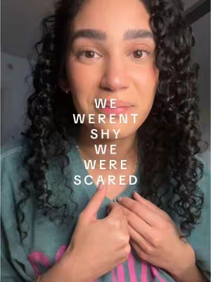 A medium shot features a person with dark, curly hair looking directly into the camera with a somber expression. Text is overlaid on the image, centered vertically with the words "WE WEREN'T SHY WE WERE SCARED" in a sans-serif font. The person's hands are near their face, with one finger pointing towards the text.