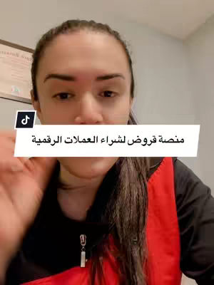 A woman with dark hair is centered in the frame, holding a phone in front of her. She is framed from the chest up. A white box with Arabic text is displayed over the bottom portion of the image. The background is a plain wall, a framed certificate is visible on the wall.