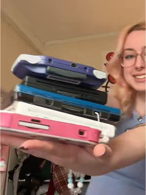 A woman with blonde hair and glasses smiles at the camera while holding a stack of handheld gaming consoles in her hand. The consoles include a purple, black, blue, white, and pink one, placed on top of each other.