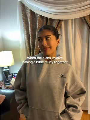 A young woman with dark hair, wearing a gray hoodie, is looking slightly to her right with a subtle smile. The background includes a curtain, a lamp, and a dark-colored table with some items on it. Text overlayed on image.