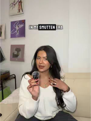 A young woman with long, dark hair is sitting in front of a white wall, holding a microphone. The text overlay says 'IN MY SMUTTEN ERA'. In the background, there are framed album covers.