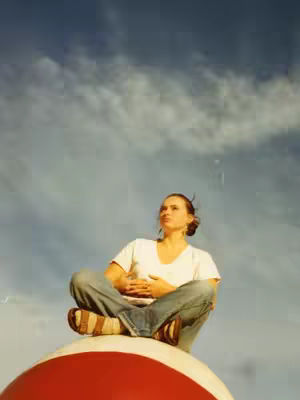 A woman sits cross-legged on top of a red and white ball with her hands clasped in front of her. She looks upwards with the cloudy blue sky as the backdrop.
