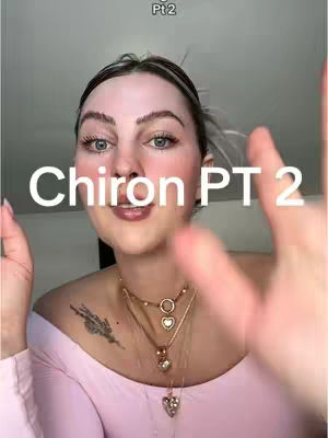 A selfie-style shot featuring a woman with fair skin, light makeup, and light brown hair with a topknot. The text overlay reads 'Chiron PT 2'. The woman wears a pink top and multiple necklaces. She is positioned in the center of the frame, with her hands near her face, partially obscuring the view.