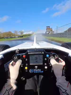 An action shot from the driver's perspective inside a race car. The steering wheel and gloved hands are in focus. Water spray obscures the road ahead, suggesting wet conditions, with a racetrack and sky in the background.