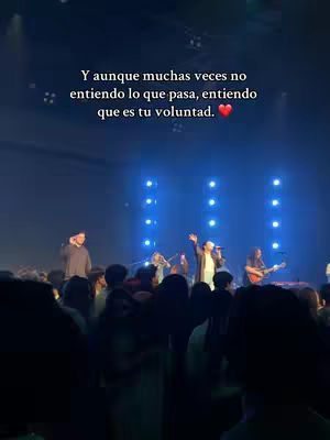 The image shows a live music performance in a large hall. A band is on stage, with several musicians playing instruments and a singer at the microphone. A large crowd of people faces the stage. Text overlayed on the top of the image reads: "Y aunque muchas veces no entiendo lo que pasa, entiendo que es tu voluntad." which translates to "And although many times I do not understand what happens, I understand that it is your will."