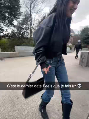 A woman in a black jacket and jeans walks towards the camera with a black purse. The background shows an outdoor setting with concrete structures and greenery, with text on the bottom as well.