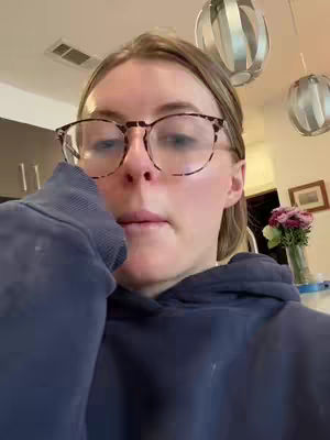 The cover image is a selfie-style shot. The main focus is on the upper body and face of a blonde woman. She is wearing round, tortoiseshell-rimmed glasses and a relaxed, navy blue hoodie. Her expression is contemplative or perhaps slightly sad. The background includes elements of a home setting, like hanging lights, a partial view of a kitchen cabinet, and a framed picture. The lighting seems to be natural and soft.