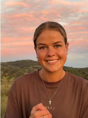 A woman with fair skin, light-colored hair pulled back, and a sincere smile poses in front of a blurred background of a landscape and a pink and orange sunset. She wears a brown t-shirt and a cross necklace, clasps her hands in front of her, and has a ring on her finger.