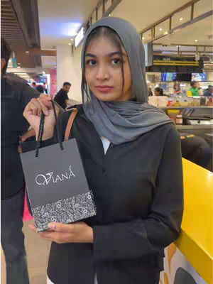 A woman wearing a hijab holds a black shopping bag in a mall setting. She is facing the camera with a neutral expression.