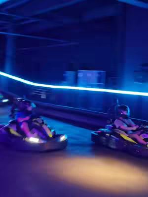 The cover image shows a dynamic shot of a go-kart track. Two go-karts are racing, each with a person wearing a helmet. The scene is lit with blue lights and appears to be indoors. Motion blur suggests speed and action.