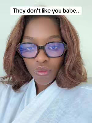 A close-up shot features a woman with brown wavy hair and black-framed glasses. The glasses reflect blue light. She has light skin, full lips, and a neutral expression. She’s wearing a white robe-like garment against a light background.