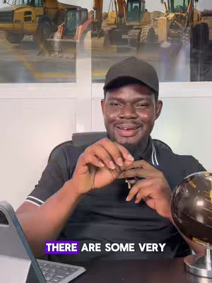 A man wearing a black cap and shirt smiles while seated with a laptop and a globe in view, with a picture of construction equipment in the background, accompanied by visible text overlay.