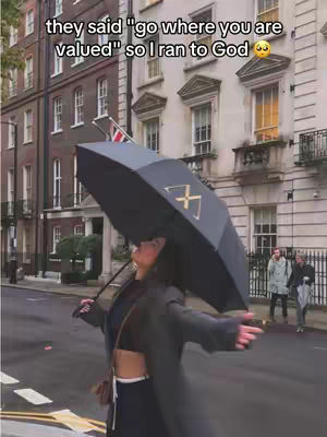 A person with long dark hair stands on a street holding a black umbrella with a gold design, looking upward with arms outstretched. They are wearing a black outfit and a long grey coat. Behind them, there are classic buildings. The sun is shining, and there is light.