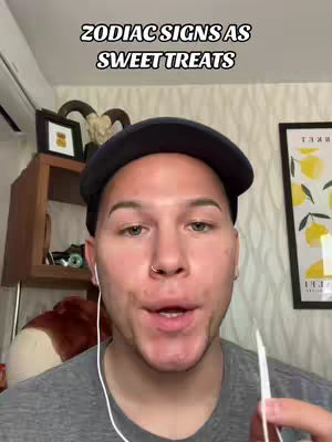 A person with fair skin and short hair wearing a black cap and grey shirt; the person is speaking into the camera, in front of a bookshelf, a framed picture, and a drawing of lemons.