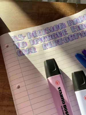 The image shows a notebook with a handwritten title about decision-making, highlighted in blue. A pink highlighter rests on the page. The setting appears to be on a wooden surface, with sunlight casting shadows across the scene.