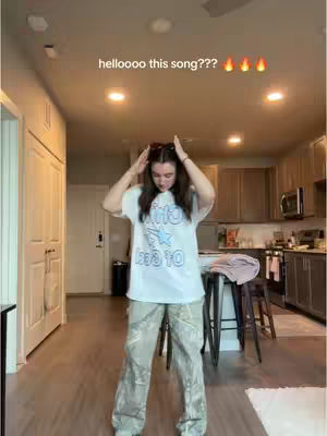 A woman poses in a home setting, wearing a white t-shirt and patterned pants. She stands in front of a kitchen background with the hook text over her head with flame emojis.