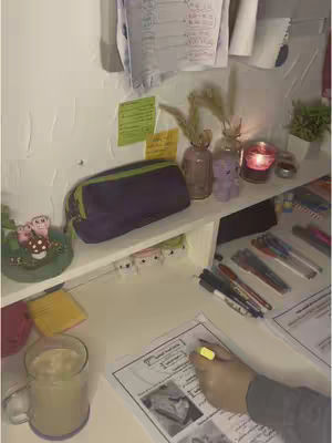 A well-lit, interior shot of a desk setup for studying. A white shelf holds various items: a purple pencil case, small pink vases, a candle, and an air freshener with a purple bear decoration. On the desk, there's a hand holding a yellow highlighter over a textbook and a mug of coffee sits next to it. The overall aesthetic is clean and organized, with a warm, cozy atmosphere.