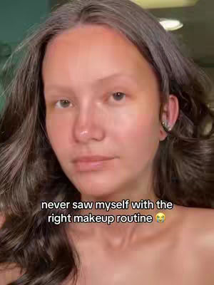 A close-up shot of a woman's face before applying makeup. She has fair skin, dark hair, and is looking towards the camera with a neutral expression. The background is blurred, with her bare shoulders visible. Text overlay at the bottom says, 'never saw myself with the right makeup routine' with an emoji.
