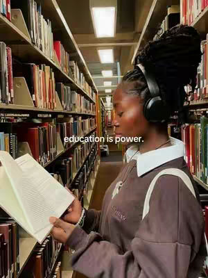 A woman with braided hair wearing headphones and a neutral-toned sweater is reading a book in a library. The background features shelves packed with books.