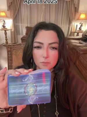 A woman with long dark hair is holding up a deck of tarot cards. The card in front is visible, with a blue and purple design and an emblem in the center. The background includes a blurry room setting, possibly a bedroom, with warm lighting from lamps and curtains.