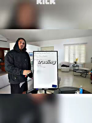 A man in a black hoodie stands in a well-lit room, pointing to a whiteboard that reads 'trading' with handwriting. The background includes a sofa, windows, and a clean, modern aesthetic. The overall composition is focused.