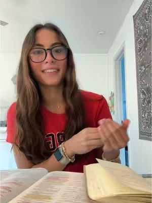 A young woman with long brown hair and glasses is seated in brightly lit room, smiling. She is wearing a red shirt and multiple bracelets and a watch. She is holding her hands up, as if to indicate the message, with open books nearby. The background consists of a wall hanging and a door.