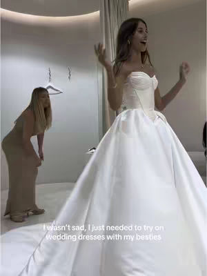 A woman in a white wedding dress stands in a bridal shop, looking up with an excited expression, arms raised. A second woman in a beige dress is in the background, also looking at the bride.