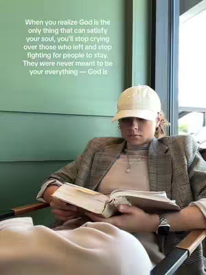 A woman in a blazer and baseball cap is seated, holding open a book. The setting appears to be an indoor space with natural lighting, suggesting a cafe or lounge area. A text overlay is present on the image.