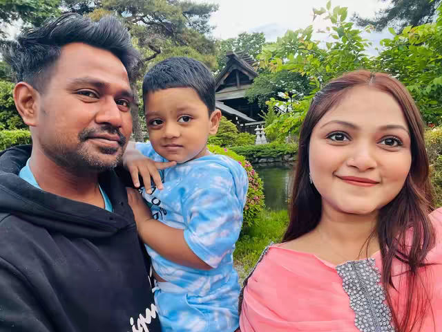 A selfie-style image featuring a man, a woman, and a young child. The man is on the left, with dark hair, a beard, and a black hooded sweatshirt. The child is in the center, held by the man the child is looking at the camera; and the woman is on the right, smiling with long hair and a pink top. A background with greenery and a Japanese-style building adds context.