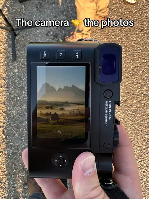 The image shows a close-up of a black camera held by a hand. On the camera's screen, a landscape with rolling hills and mountains is visible. The text overlay reads 'The camera 🤝 the photos'. Parts of an unseen person's outfit are visible in the background.