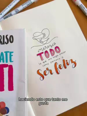 A colorful image features a notebook with hand-lettered text and drawings. The text 'Mereces Todo' in pink and orange, along with 'Ser Feliz' and other phrases, is displayed with decorative elements. A second notebook is partially visible on the left side of the image, and various markers are placed around the main notebook, all on a wooden surface.