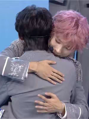 Two men embrace in a close-up shot. One has his back to the camera, wearing a grey suit. The other, with pink hair, is leaning his head on the first man's shoulder and embracing him. The background has a blurred blue tone.