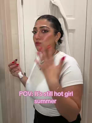 A woman with dark hair and makeup stands in front of a mirror, with red nail polish and a white shirt. Text is overlaid, and the composition is a selfie.