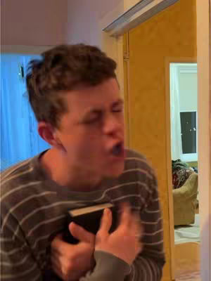 A man with short brown hair, wearing a striped shirt, is shown from the chest up, his face is expressive, eyes closed and mouth open as he is gesturing. He is holding a book to his chest.