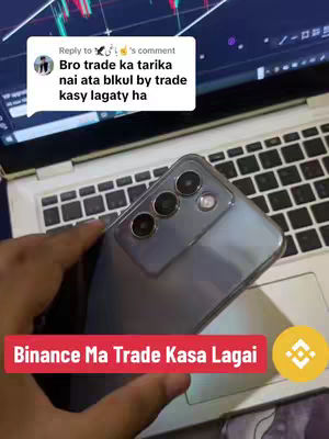 The cover image showcases a close-up view, focusing on a phone held in a hand. The phone is positioned over an open laptop. A text overlay in a red box at the bottom of the image reads 'Binance Ma Trade Kasa Lagai.' A comment above the phone is superimposed from a reply to the user. The laptops displays trading charts. The background is a blurred view of a room. The overall composition is a blend of technology and text overlay.