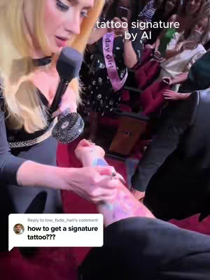 A woman with blonde hair speaking into a microphone while holding a tattoo gun near a leg, surrounded by other people. The scene is lit with warm lighting.