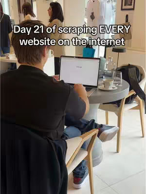 A man is sitting at a table, typing on a laptop. The laptop screen displays a Google search. There are other people in the background, and the setting appears to be a cafe. Text overlayed on the image reads: \"Day 21 of scraping EVERY website on the internet\".