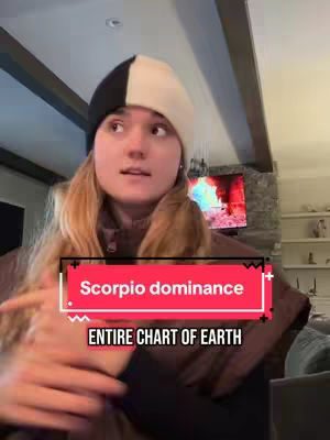 A young woman with long blonde hair is wearing a black and white beanie and a brown jacket. She is centered in the frame, looking slightly off-camera with a neutral expression. A red rectangular graphic with the words "Scorpio dominance" and "ENTIRE CHART OF EARTH" is superimposed over her chest. Behind her is a blurred background with a fire place and a television. The overall composition is lit with soft, natural light.