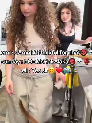 The image shows two young women in a kitchen setting. The woman in the foreground has long, curly hair and is wearing a white top and gray sweatpants. The woman appears to be holding something. A second woman is behind her, wearing a black top and jeans. Overlaid text includes conversational snippets and emojis.