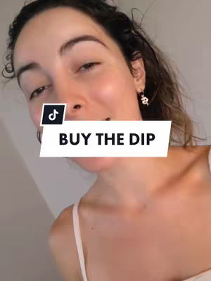 A selfie shows a woman with dark hair, looking directly at the camera and smiling slightly. She's wearing a white top. A white banner with the text 'BUY THE DIP' is positioned over her mouth. The background is blurred and minimalistic.