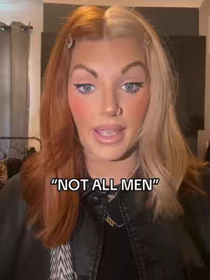 A close-up shot of a person with a split hairstyle, half blonde and half auburn. The person is looking at the camera with an open mouth, wearing black eyeliner and a black top. Text overlay at the bottom reads "NOT ALL MEN."