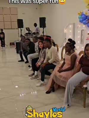 A group of people are seated, likely indoors for a party. There is a DJ set up in the background and decorations, suggesting a birthday party. The hook text on the top announces that the game played at the party was fun.